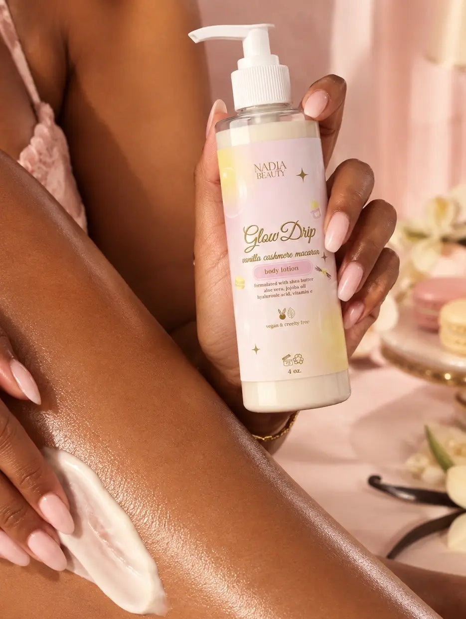A person holding a bottle of Nadia Beauty Glow Deep Body Butter while applying a white cream lotion to their leg. The bottle is cream-colored with a white pump and gold lettering, set against a soft pink background.