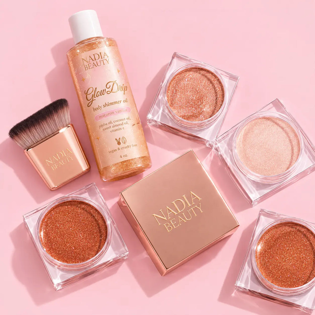 Nadia Beauty makeup set arranged on a pink background, including a Glow Drop body shimmer oil bottle, a rose-gold makeup brush, and four square jars of shimmering face and body powders in copper, peach, and champagne tones.