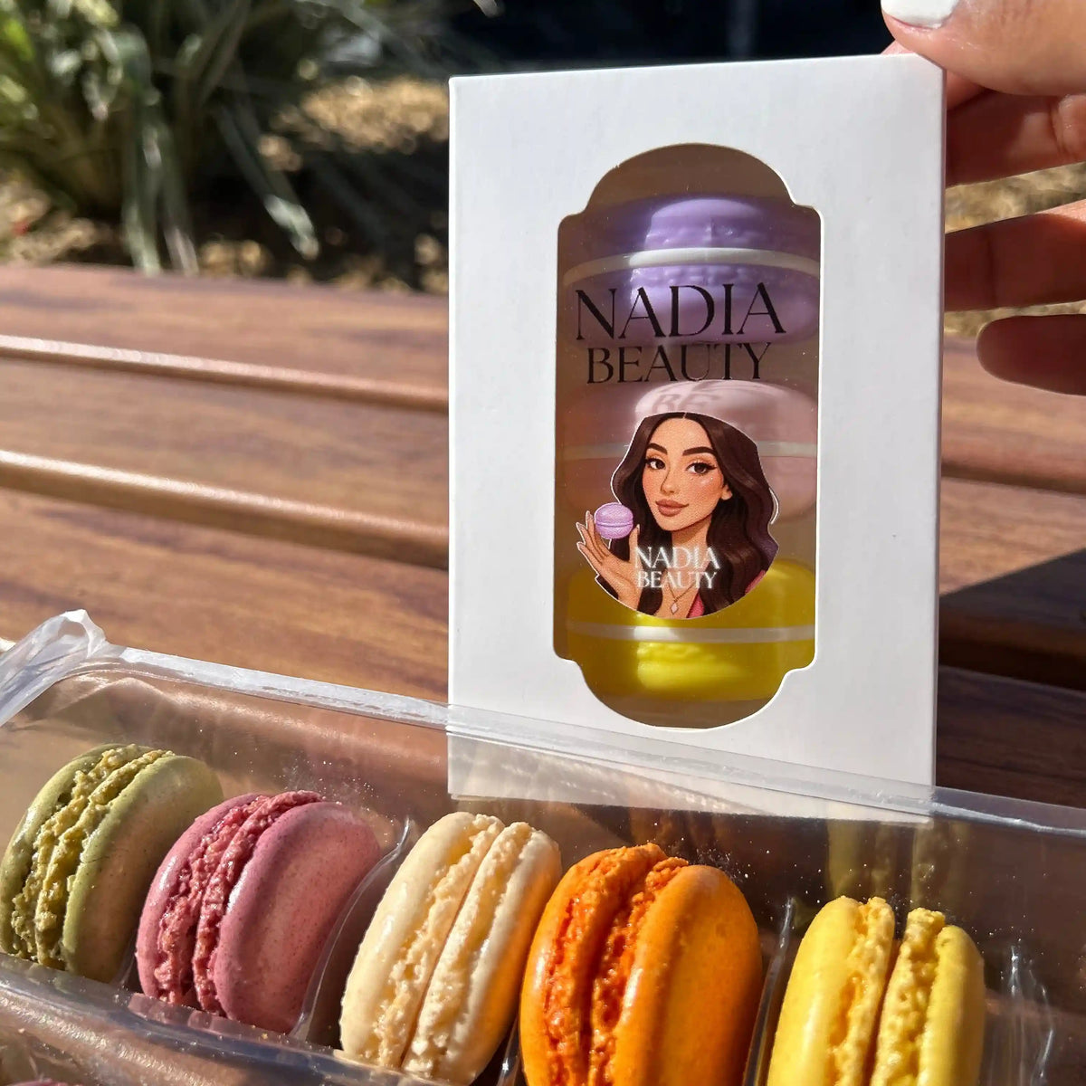 A hand holds a white box labeled 'NADIA BEAUTY' in front of a clear plastic tray of colorful macarons on a wooden outdoor table.
