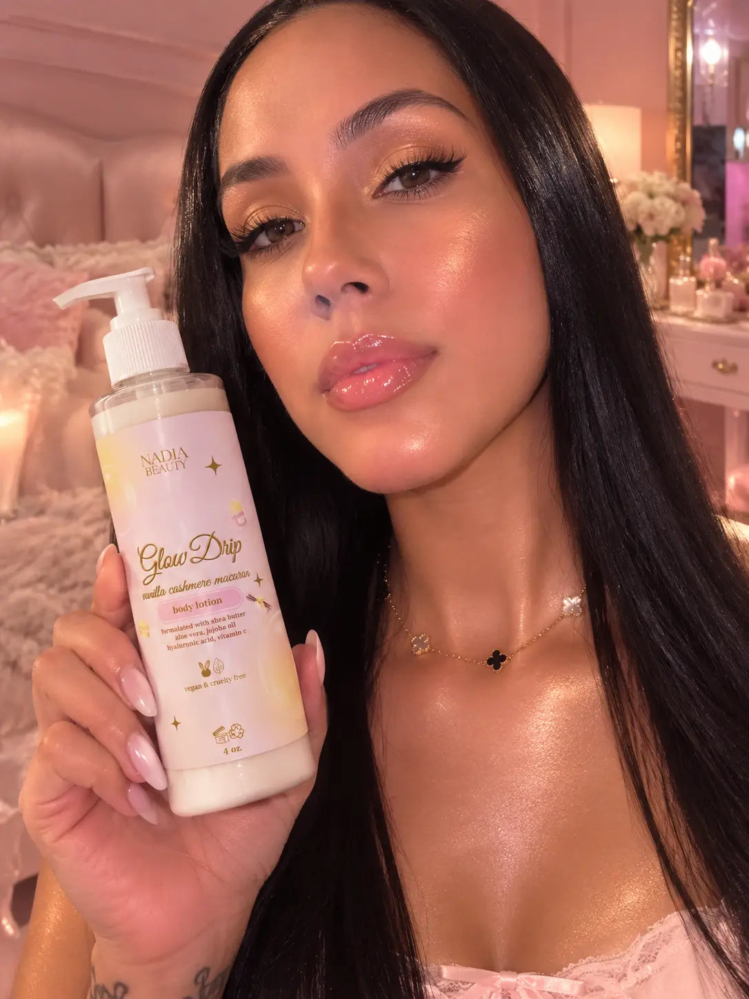 A woman holds a bottle of NADIA Beauty Glow Dip body lotion in a pink bedroom setting. The bottle is white with gold lettering and a white pump dispenser.