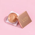 A small round cosmetic pot filled with shimmery gold makeup, shown next to its square rose-gold lid printed with the brand name 'NADIA BEAUTY' in gold, set against a solid pink background.