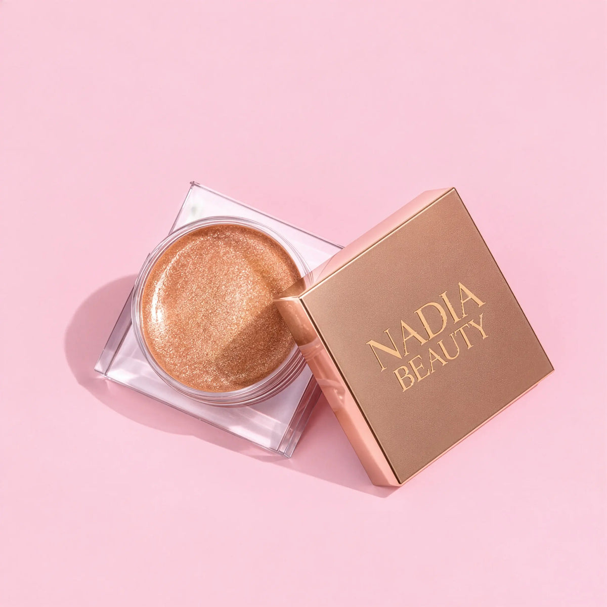 A small round cosmetic pot filled with shimmery gold makeup, shown next to its square rose-gold lid printed with the brand name 'NADIA BEAUTY' in gold, set against a solid pink background.
