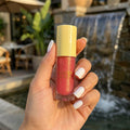 Hand holding a rosy pink lip gloss with gold cap, blurred outdoor background