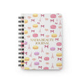 A spiral-bound notebook with a white cover decorated with pink and yellow macarons, pink bows, Eiffel Towers, and small pink teacups. The center of the cover features the gold text 'NADIA BEAUTY JOURNAL'.