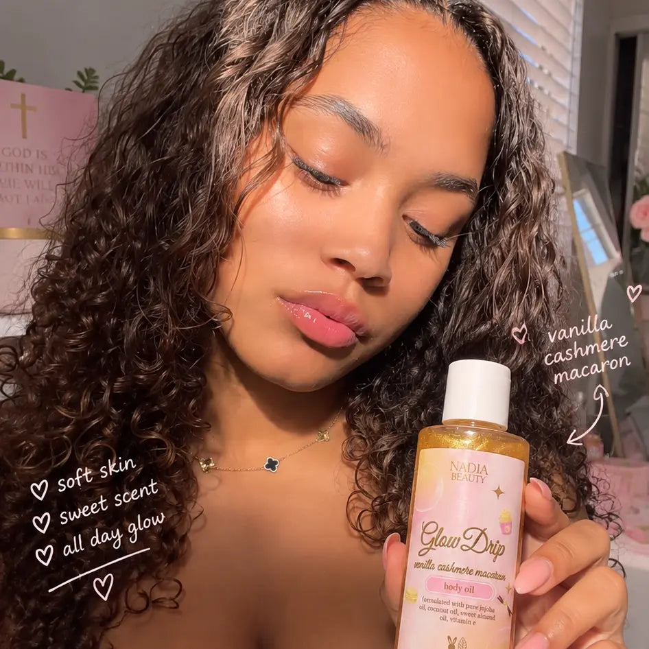 A woman with long, curly brown hair holds a bottle of NAIA BEAUTY Glow Dip vanilla cashmere macaron lavender oil. The image features handwritten text overlays highlighting the soft skin, sweet scent, and all-day glow, with a cozy room background.