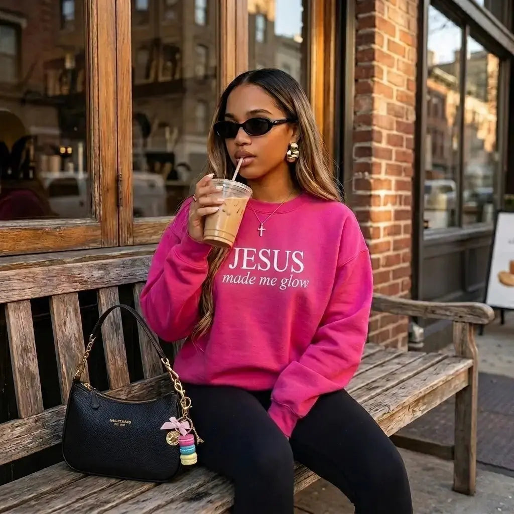 Woman in pink 'Jesus made me glow' sweatshirt sitting on a bench with a drink, wearing sunglasses.